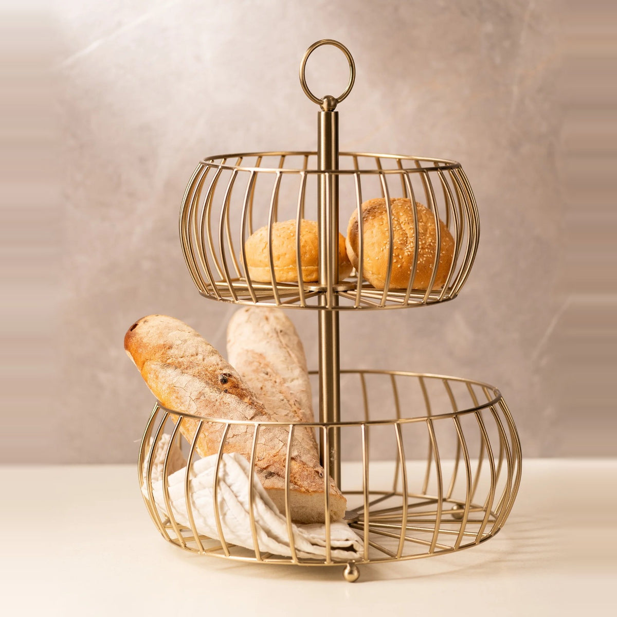 Two-tier gold wire fruit basket holding bread rolls and a baguette