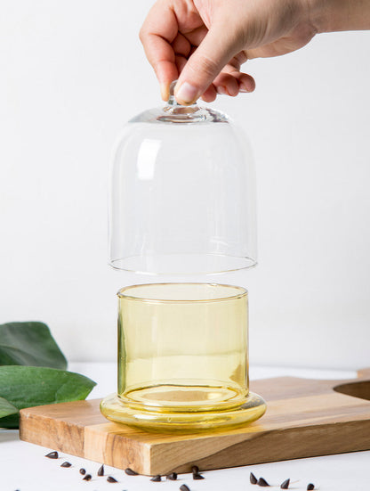 Hand lifting the clear cloche lid off a honey-yellow glass bell jar base