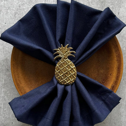 Gold pineapple napkin ring holding a navy linen napkin on a wooden plate