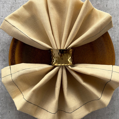 Hammered gold napkin ring gathering a cream linen napkin on a wooden plate