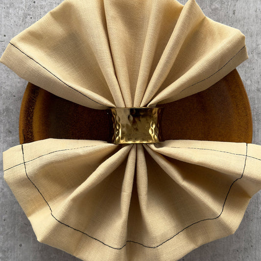 Hammered gold napkin ring gathering a cream linen napkin on a wooden plate