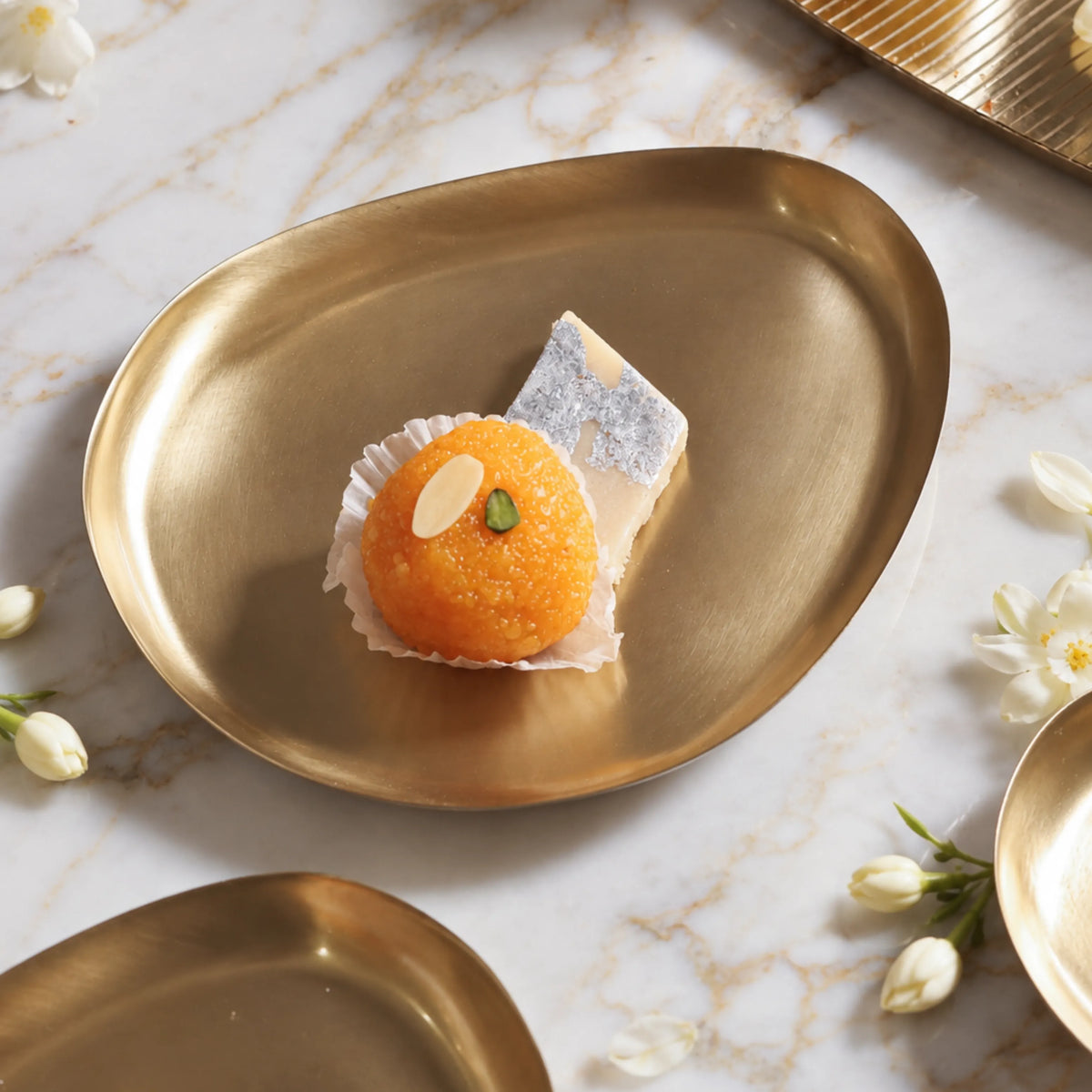 Pebble-shaped brass dessert plate holding a laddoo and a square of kaju katli