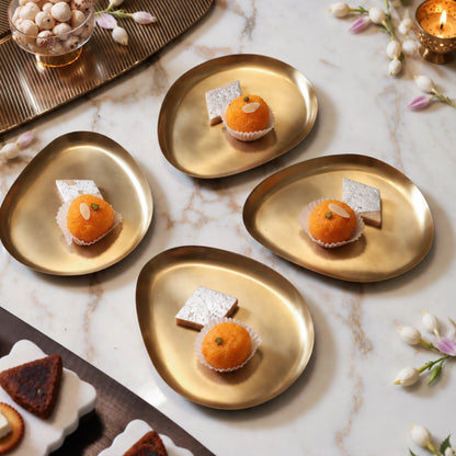 Four pebble-shaped brass snack plates with laddoo and kaju katli on a white marble table