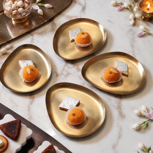 Four pebble-shaped brass snack plates with laddoo and kaju katli on a white marble table